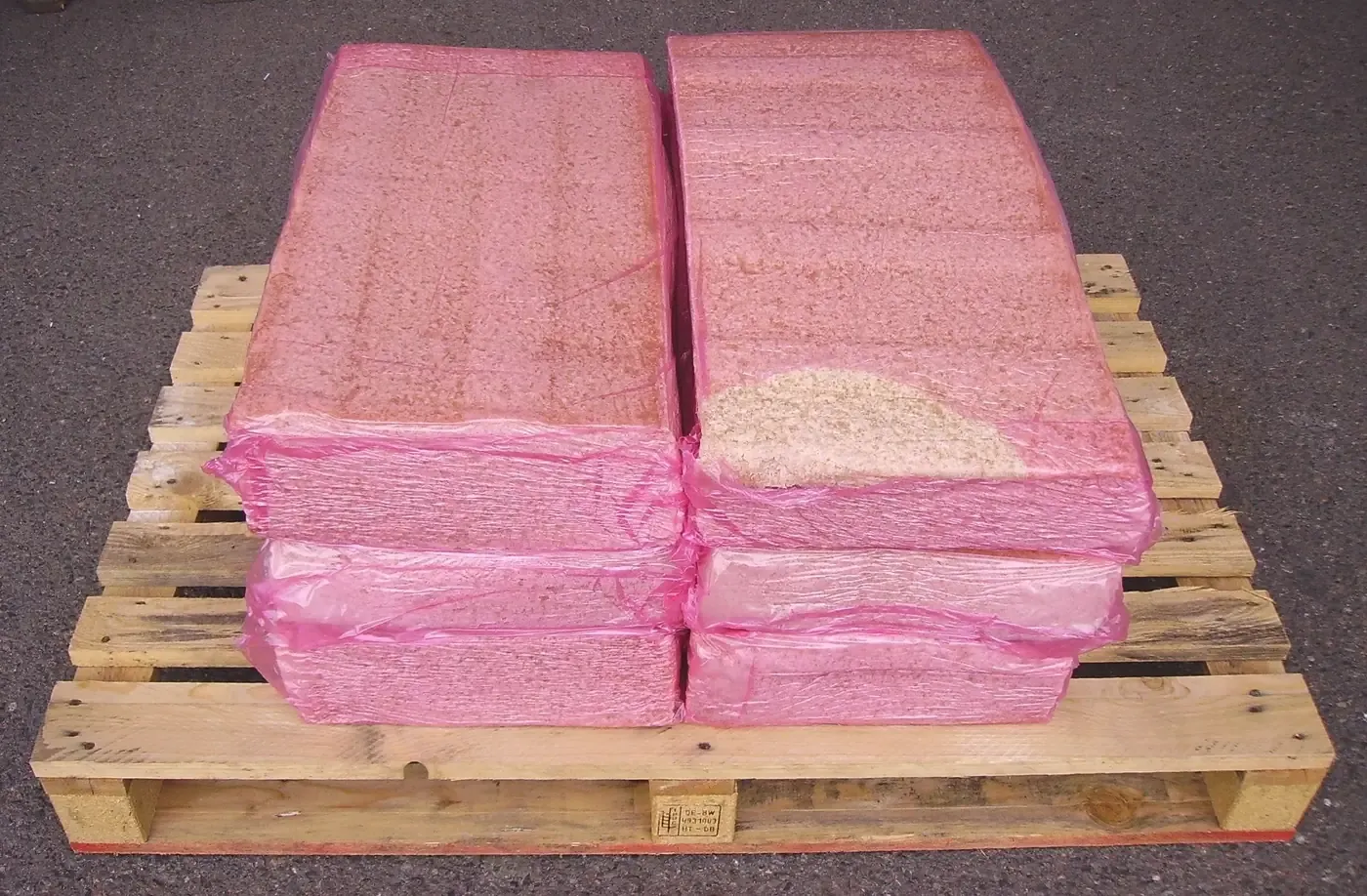 Six plastic-wrapped rectangular bales of pale tan rubber crumb, stacked two high on a wooden pallet, the wrapping torn open at one corner to show the granular material inside.
