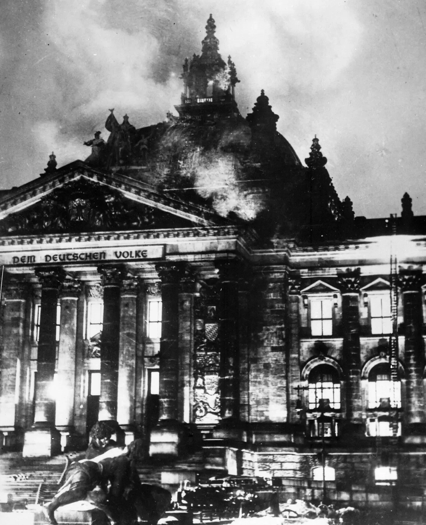 The Reichstag building at night, its great dome and colonnade lit from within by fire, smoke pouring from the roof.