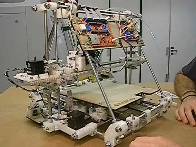 A homemade desktop 3D printer built from metal threaded rods and 3D-printed white plastic brackets, with circuit boards and wiring visible on top and a build platform below.