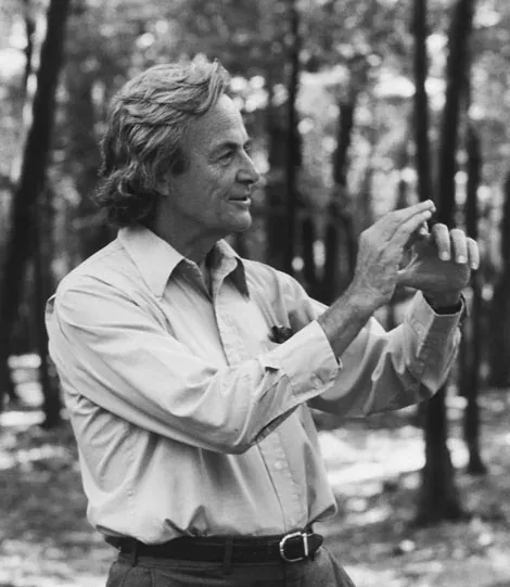 A grey-haired man in a light shirt, seen in profile outdoors among trees, holding one hand raised as if gesturing mid-explanation.