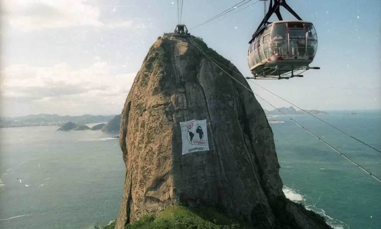 An aerial photograph of Sugarloaf Mountain in Rio de Janeiro, with a cable car passing in the foreground and a large white banner reading 'SOLD / VENDIDO' with a Greenpeace logo hung across the rock face.