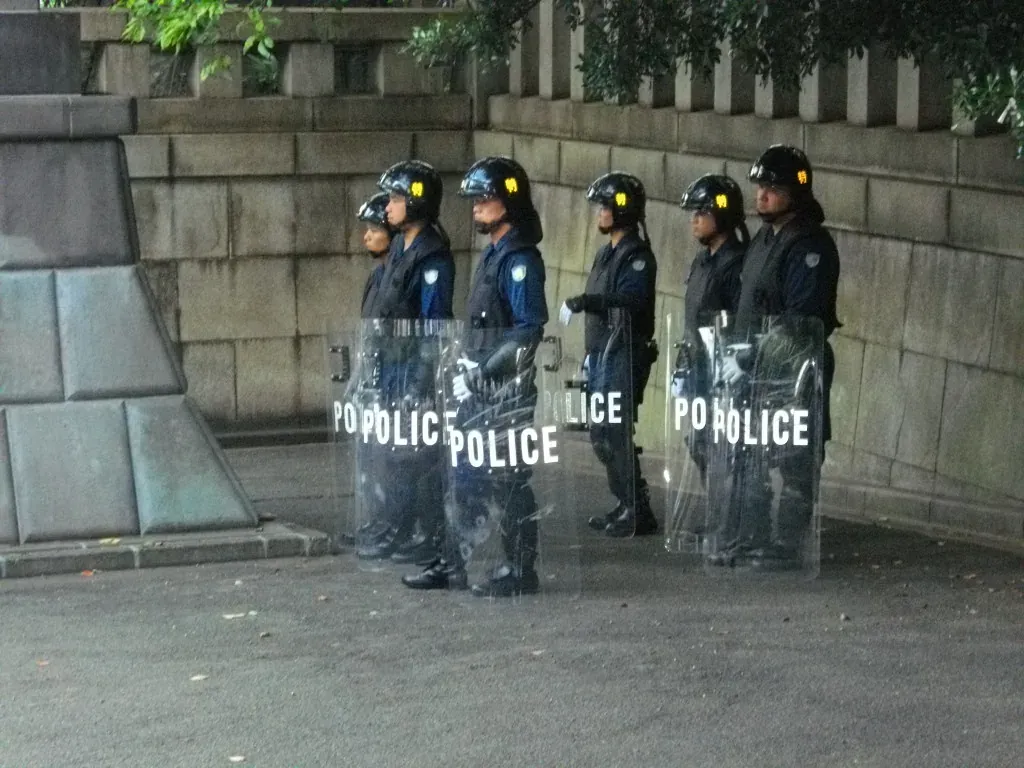 Six police officers in helmets and dark uniforms stand in a line against a stone wall, each holding a large transparent shield printed with the word POLICE in white capital letters.