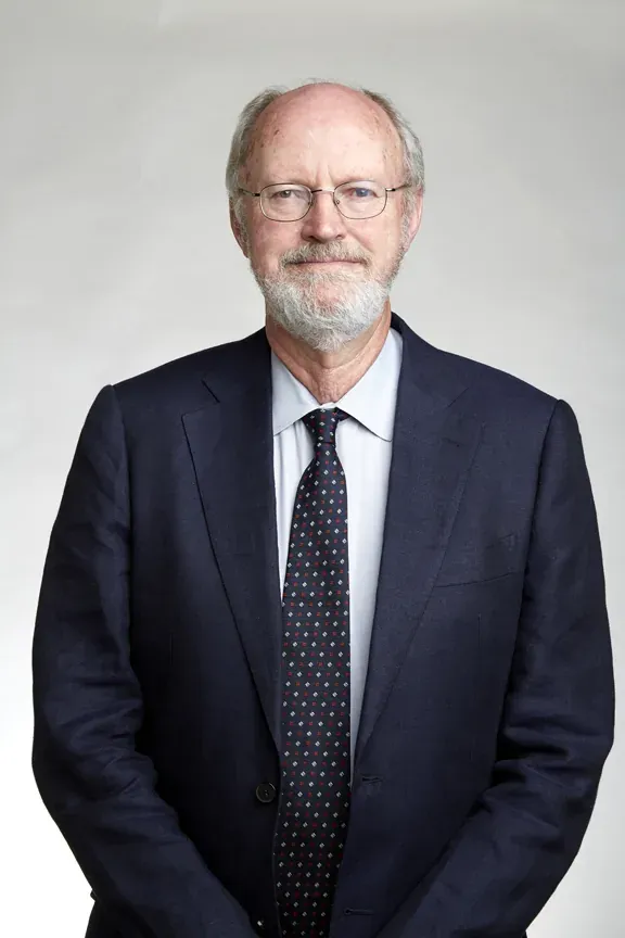 A formal portrait of an older man with a grey beard and wire-rimmed glasses, wearing a dark suit and patterned tie, against a plain grey background.