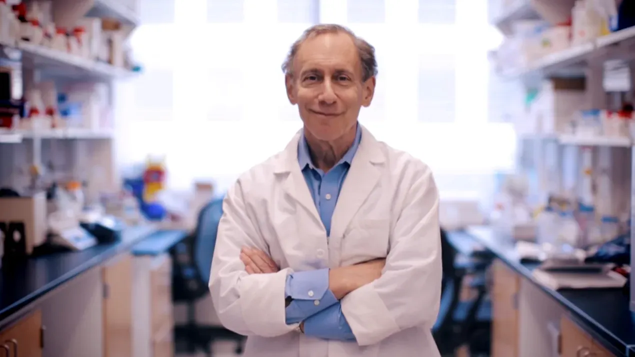 An older man with grey hair, wearing a white lab coat over a blue shirt, standing with arms crossed in a laboratory lined with shelves of reagent bottles and equipment.