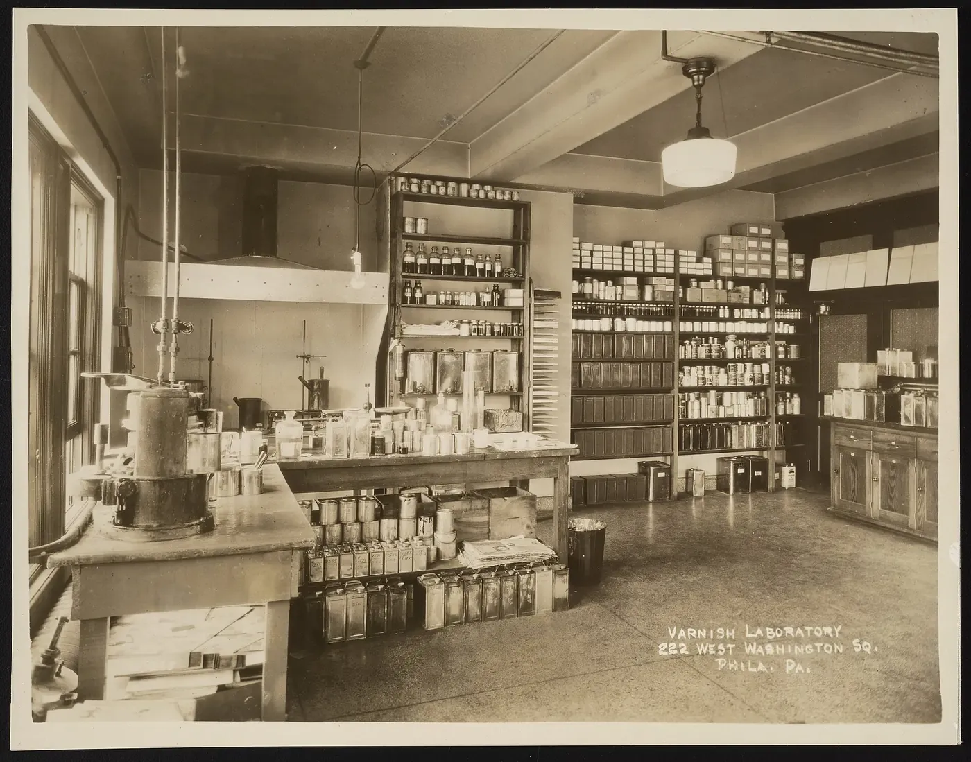A vintage photograph of a chemistry laboratory workbench crowded with cans, bottles, and glass apparatus, with shelves of labelled tins along the back wall, captioned 'Varnish Laboratory, 222 West Washington Sq., Phila. Pa.'