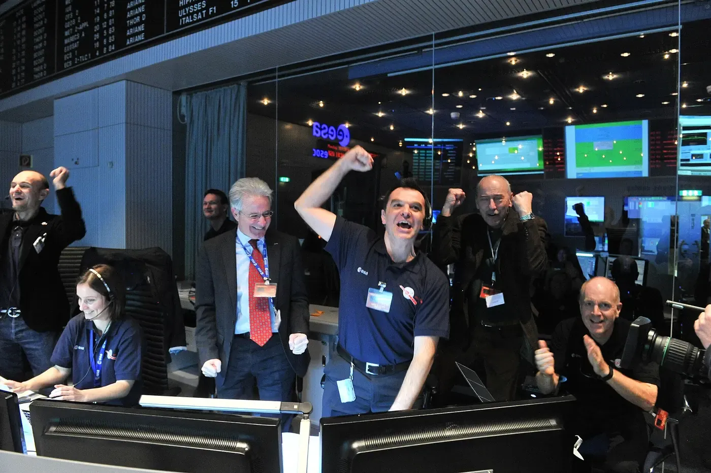 A group of engineers in a space mission control room, several with fists raised and mouths open in celebration, standing at rows of monitors below screens reading 'ESA'.