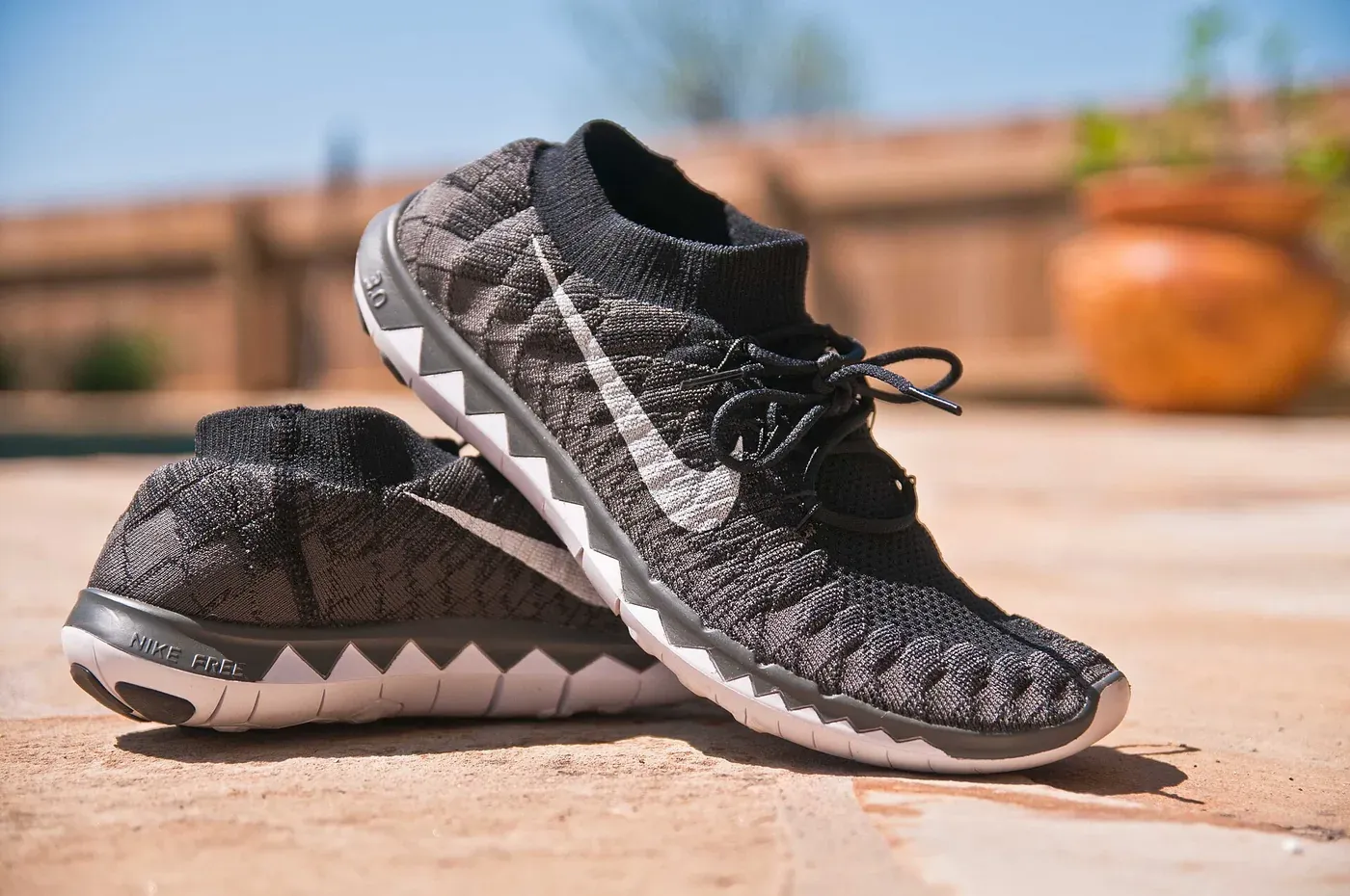 A pair of black-and-grey knit running shoes with white ridged outsoles, standing on a sunlit paved surface.