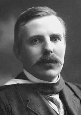 A formal studio portrait of a mustached man in a dark suit and high collar, looking directly at the camera.