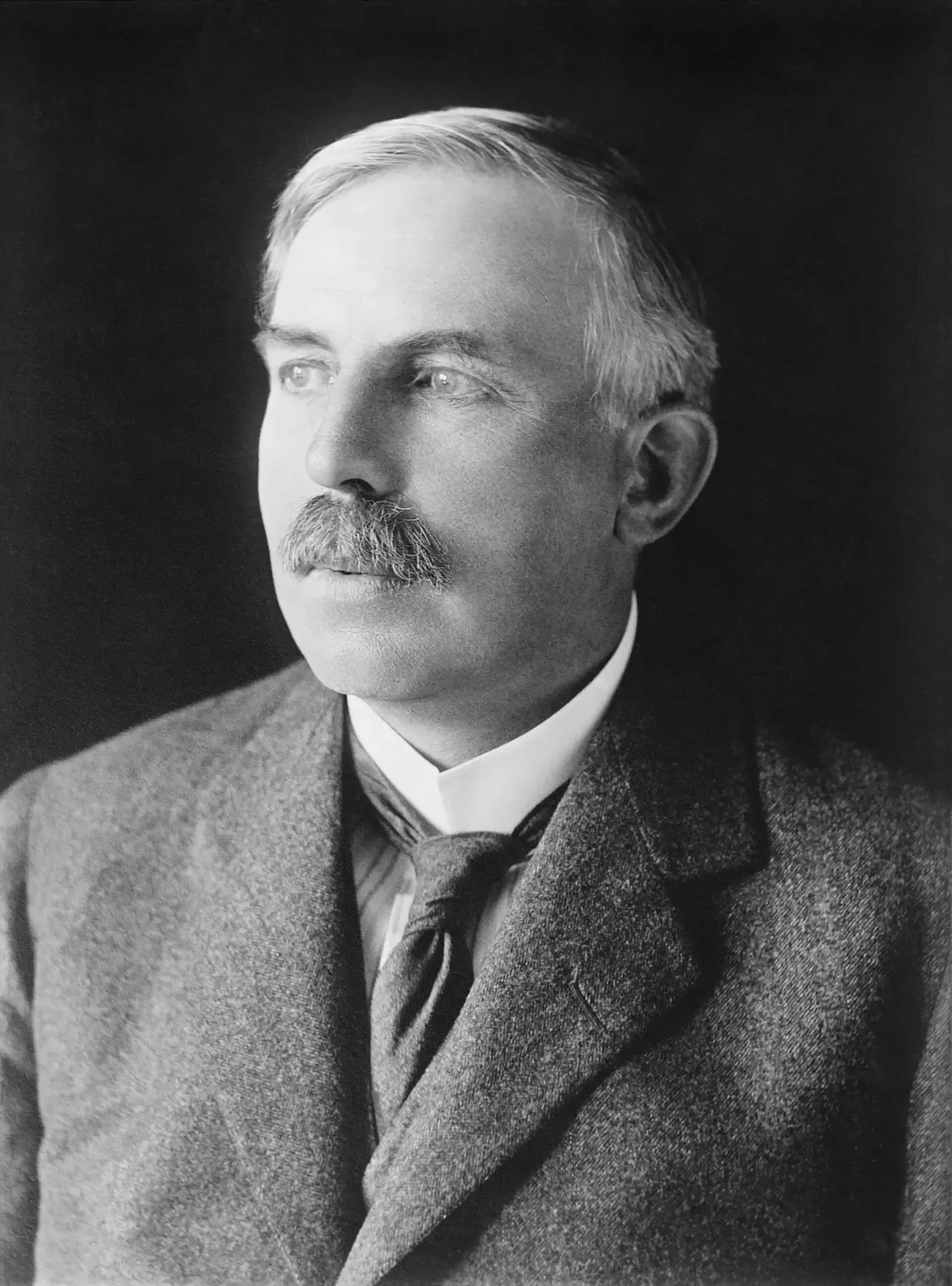 A formal studio portrait of a balding man with a bushy grey moustache, in a tweed suit and dark tie, looking off to one side.