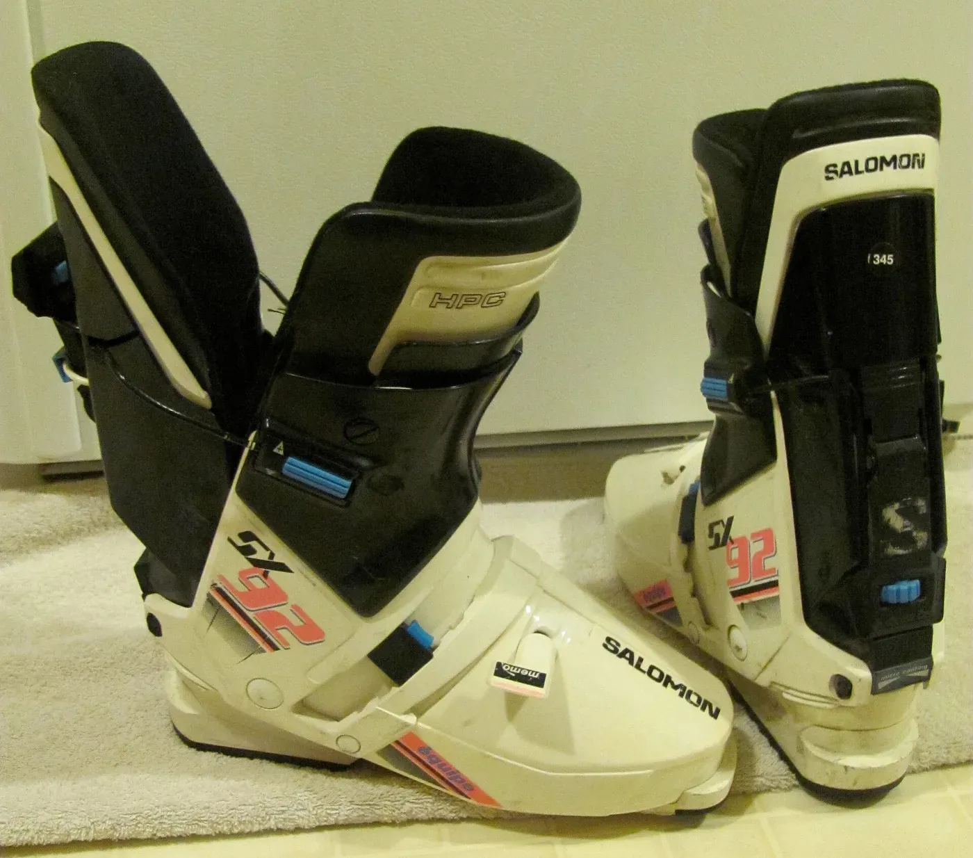 A pair of white and black rear-entry ski boots, one shown open with its rear cuff swung back, the other closed, photographed against a plain wall.