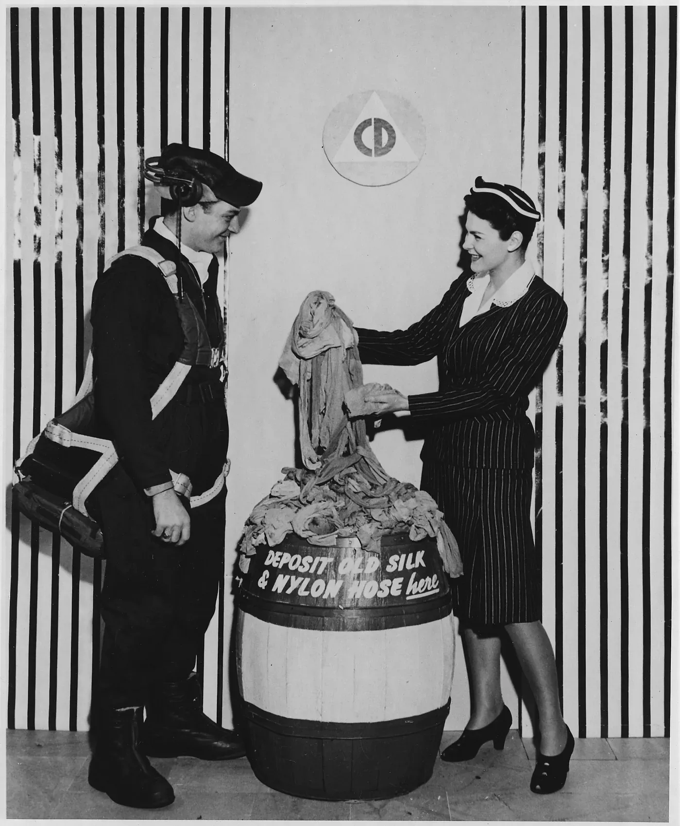 A black-and-white photograph of a woman in a pinstriped suit dropping a pair of stockings into a wooden barrel stencilled 'DEPOSIT OLD SILK & NYLON HOSE HERE', while a man in flight gear and a headset looks on.