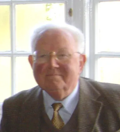 An elderly man with white hair and glasses, in a tweed jacket, smiling, photographed indoors near a window.
