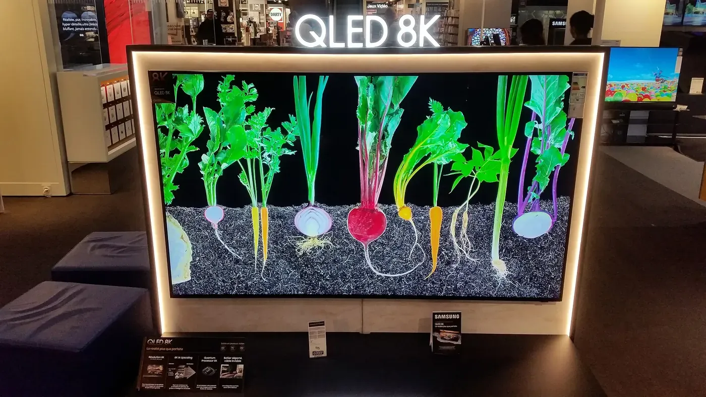 A large flat-screen television on a showroom floor, illuminated 'QLED 8K' lettering above it, displaying a vivid, saturated image of root vegetables against a black background.