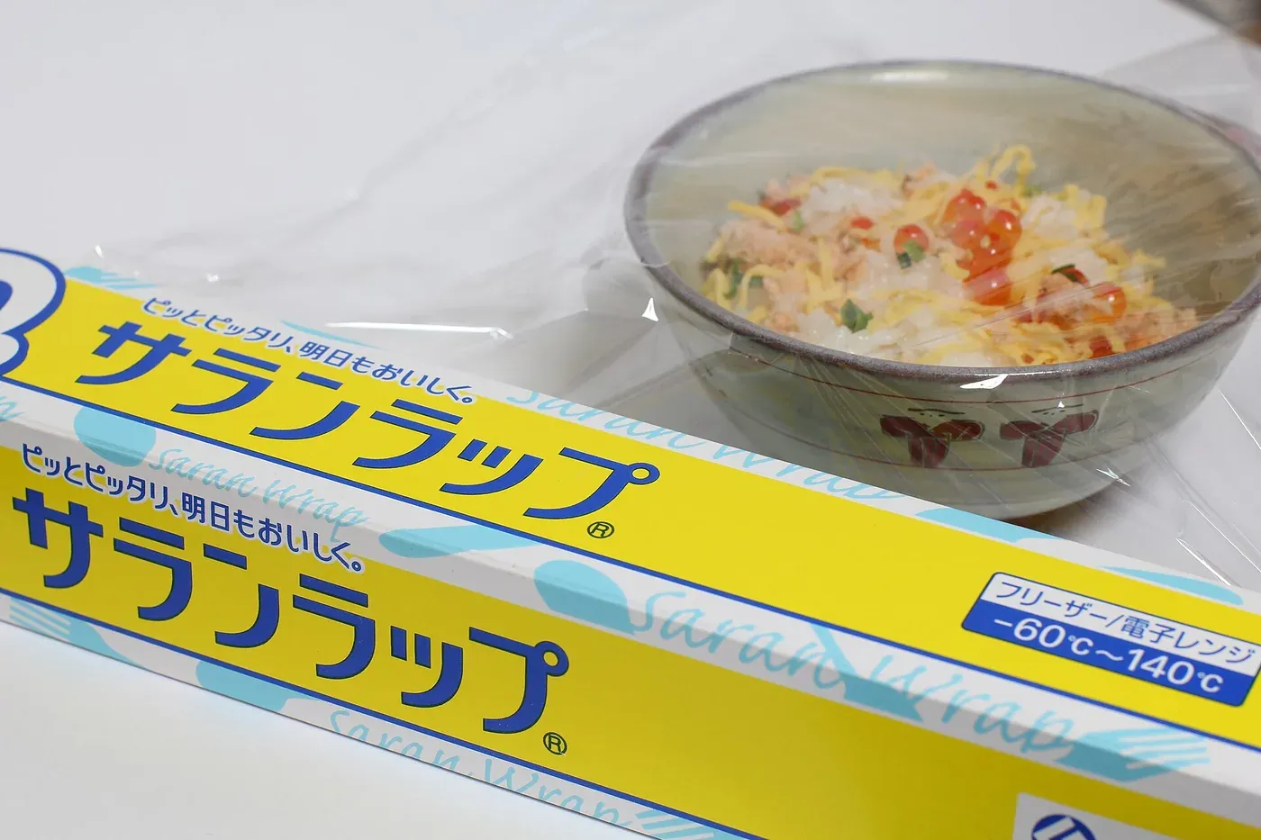 A yellow box of Saran Wrap cling film, its plastic film partly unrolled and stretched over a bowl of food on a table.