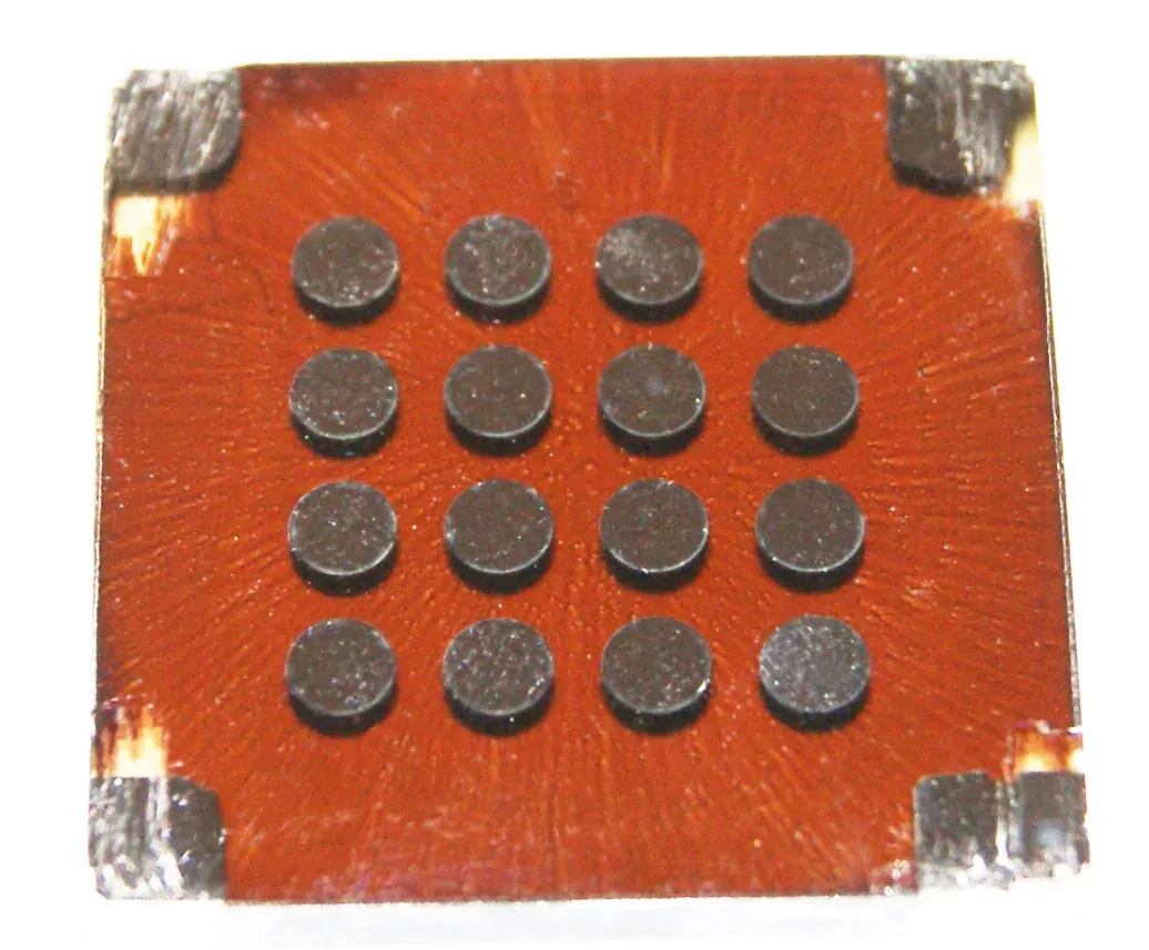 A square reddish-brown photovoltaic device on a white background, its surface patterned in radiating streaks with four rows of four dark circular metal electrode contacts.