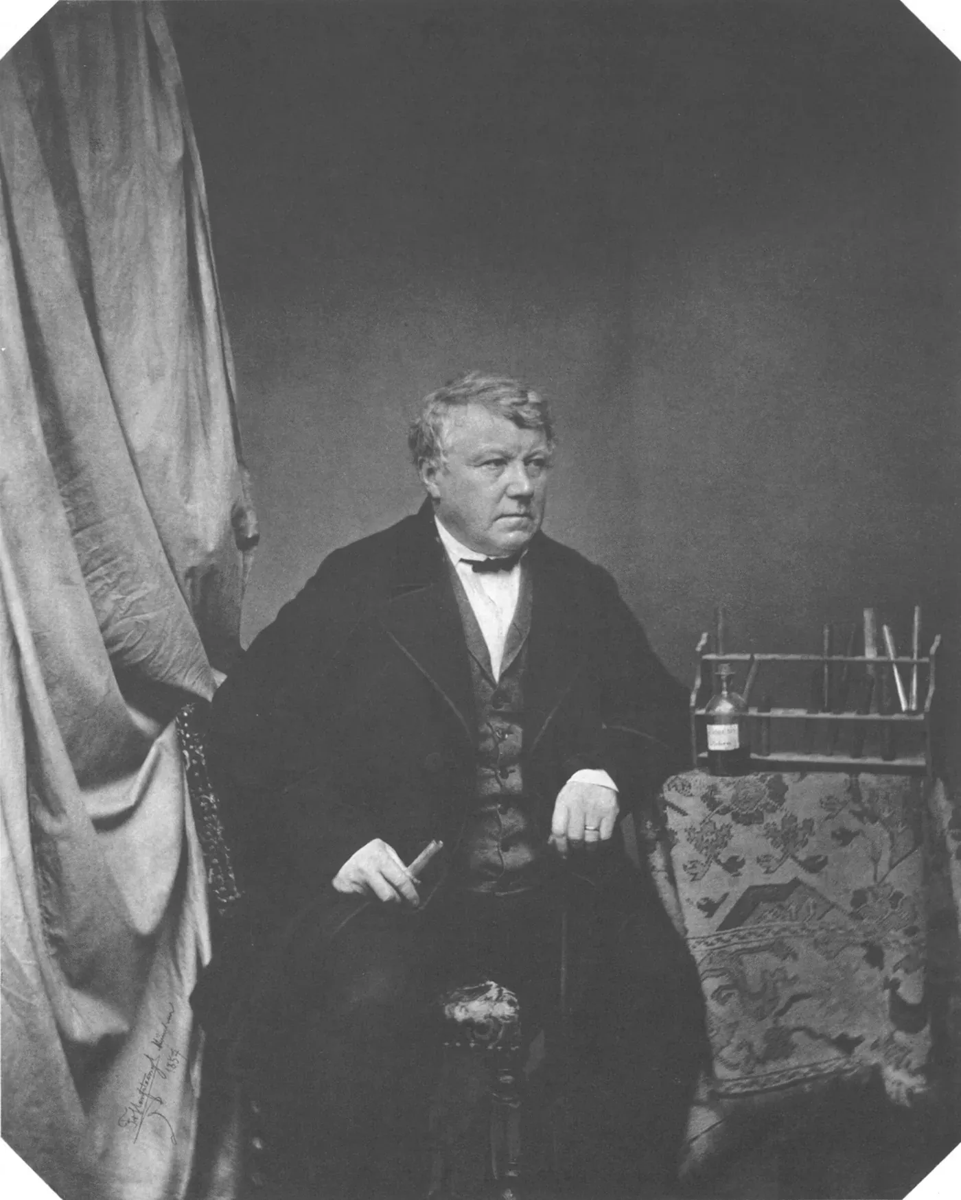 A seated studio portrait of an older man in a dark coat and waistcoat, a cigar in one hand, with a rack of chemistry reagent bottles on a table beside him.