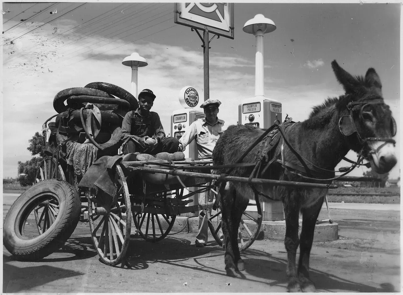 A donkey pulling a two-wheeled cart heaped with old tires past a filling station, with two men seated among the load.