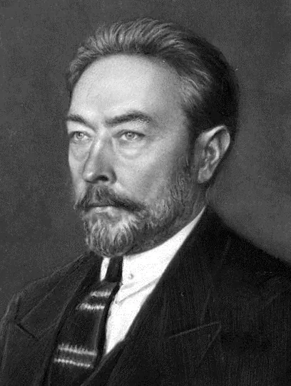 A black-and-white studio portrait of a bearded man with short combed-back hair, wearing a dark suit and striped tie, photographed from the shoulders up.