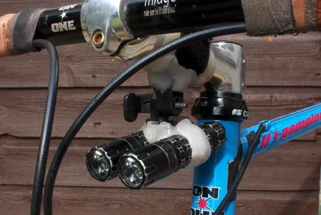 A close-up of a bicycle handlebar stem where two flashlight torches are clamped in place by a lump of hand-shaped white plastic moulded around the metal fittings.