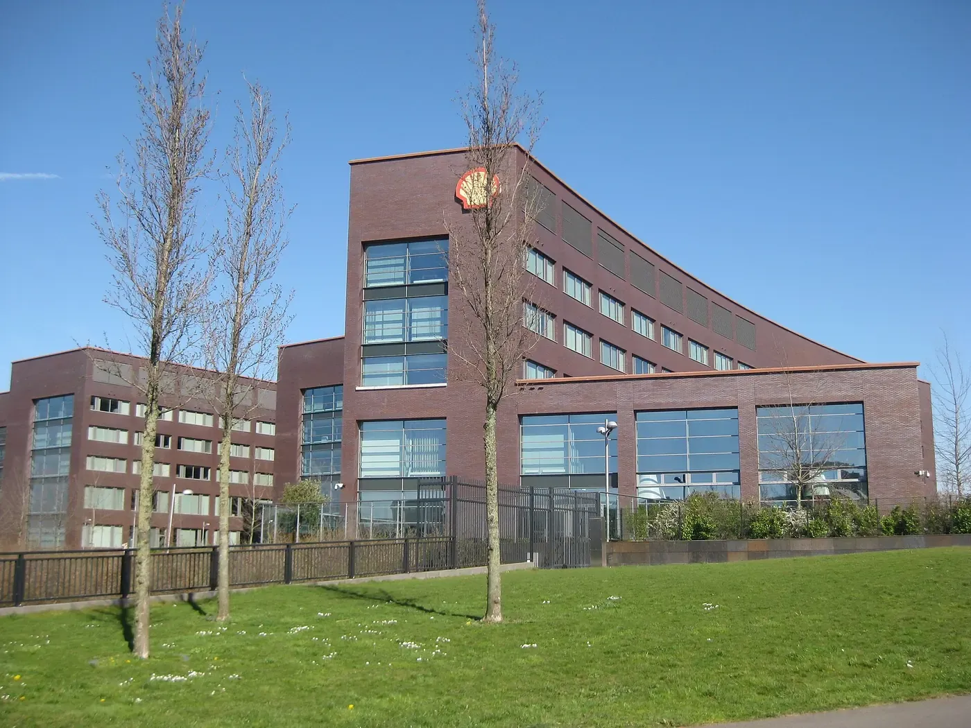 A modern multi-storey brick office and laboratory building with a large Shell shell-logo mounted near its roofline, seen across a lawn planted with young trees.