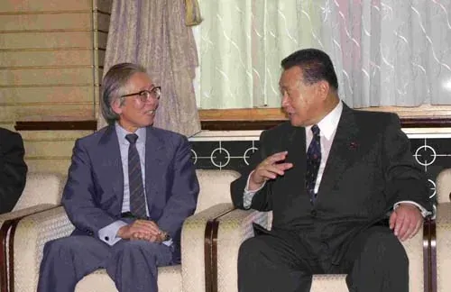 Two men in suits sitting in armchairs in conversation; the man on the left wears glasses and holds his hands together, and the man on the right gestures as he speaks.