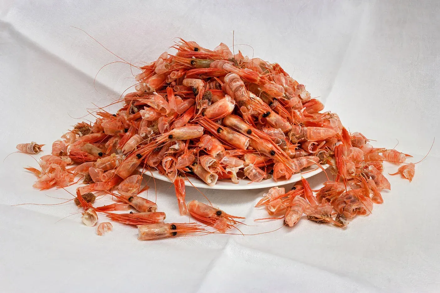A large heap of pink-orange shrimp shells and heads piled on a white plate, set on a white cloth.