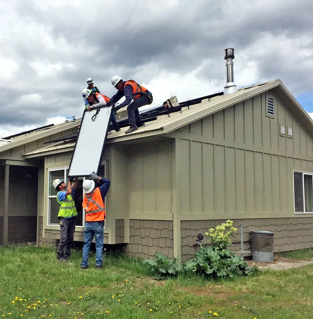 Four workers in hard hats and safety vests lift a solar panel onto the roof of a house under a cloudy sky.