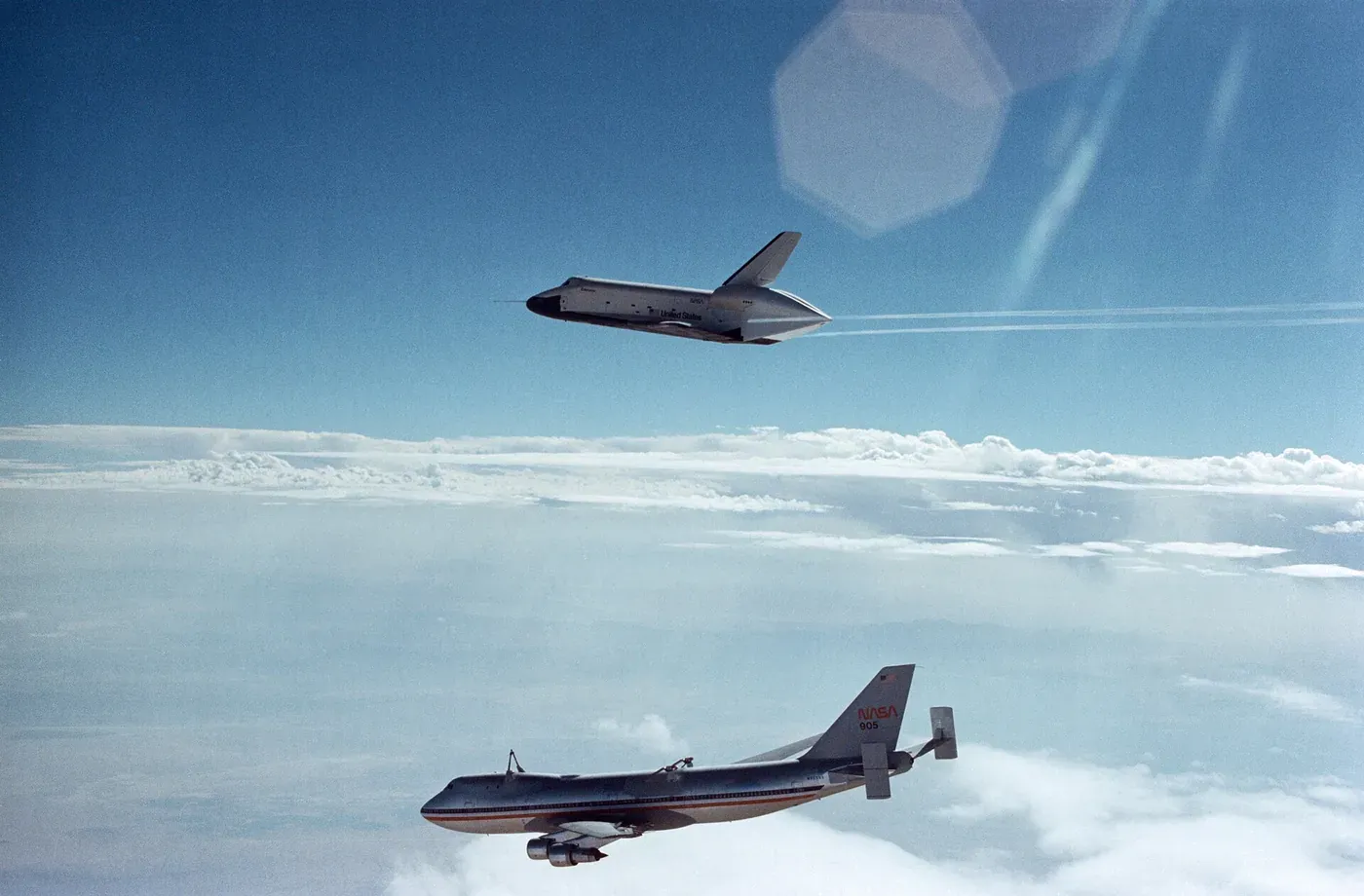 The wingless-looking Space Shuttle prototype Enterprise gliding above cloud cover, having just separated from the NASA 747 carrier aircraft flying below it.
