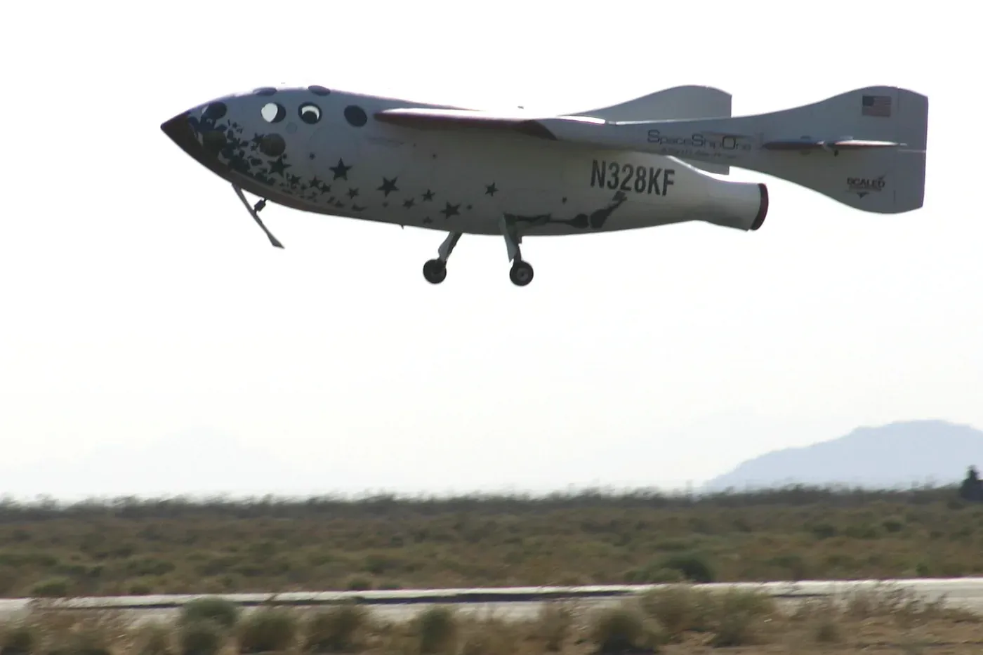 A small white rocket-powered aircraft with a bulbous nose, twin tail booms and stubby wings, coming in to land over scrubland with its landing gear extended and its wings in the flat, unfeathered position.