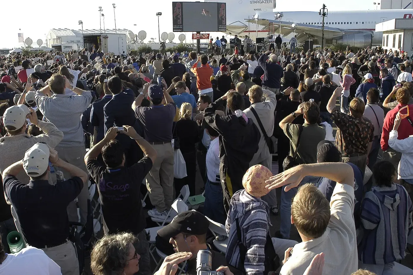 A dense crowd at an airfield, most people holding up cameras or shading their eyes as they watch the sky, with a jumbo jet-sized carrier aircraft and a large video screen behind them.