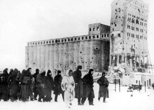 A long column of German prisoners of war, heavily bundled against the cold, walking past the bombed and shell-pocked ruins of a large grain elevator building in the snow.