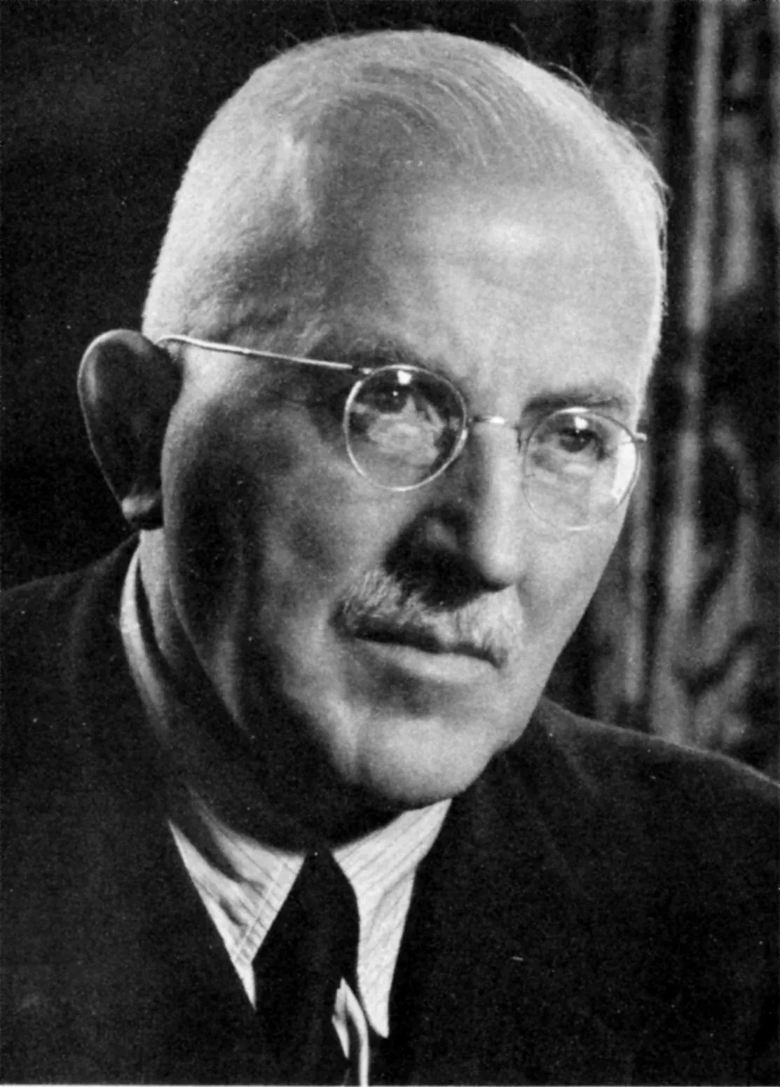 A balding man in his sixties wearing round wire-rimmed glasses and a dark suit with a striped tie, photographed in three-quarter profile.