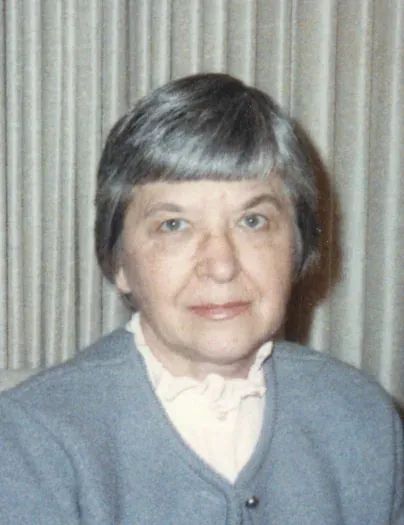 An informal head-and-shoulders portrait of an older woman with short grey hair, wearing a grey cardigan over a ruffled blouse, photographed against striped curtains.