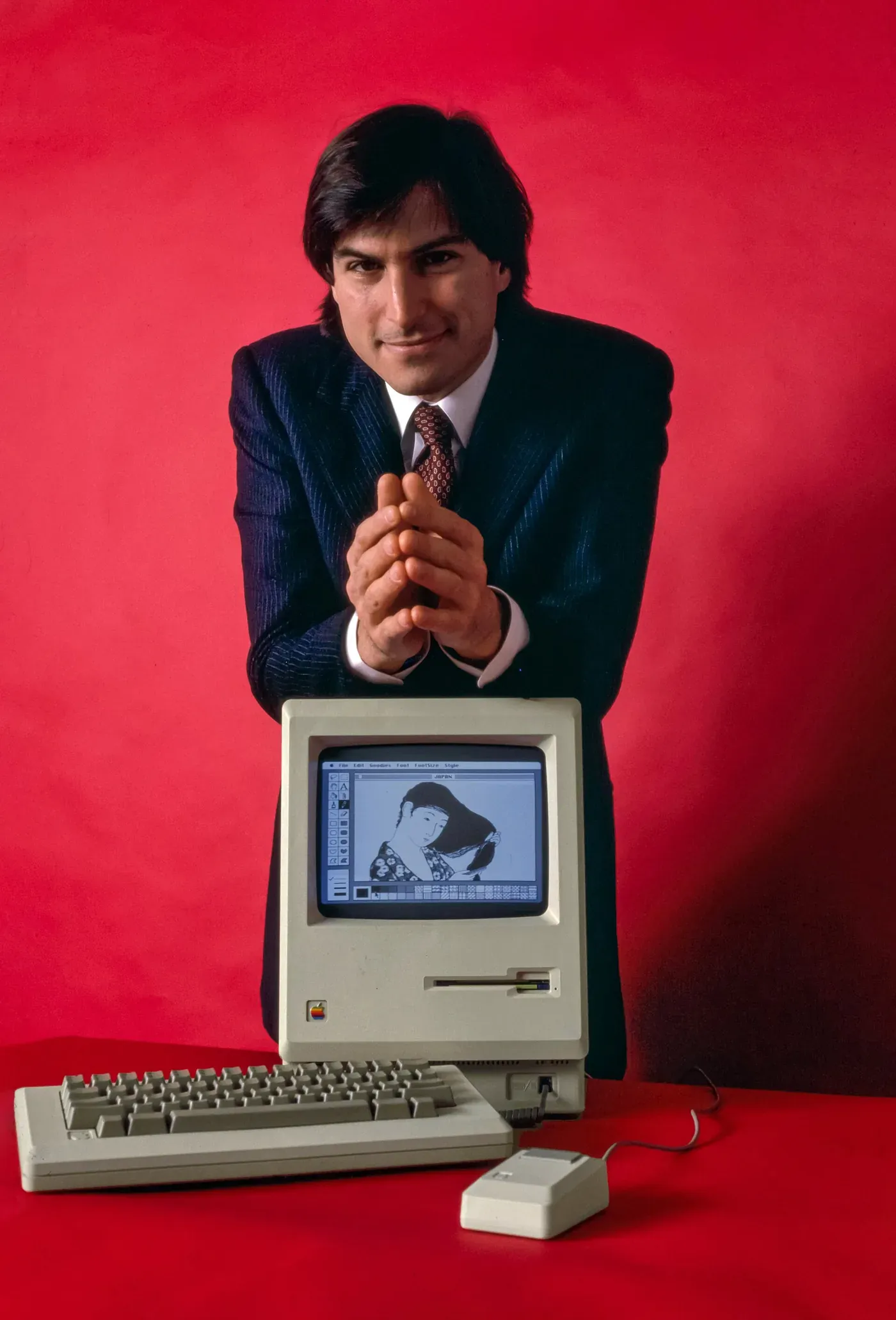 Steve Jobs, in a dark suit, leans over an original beige Macintosh computer with hands clasped, its screen displaying a line drawing of a woman combing her hair, against a plain red background.