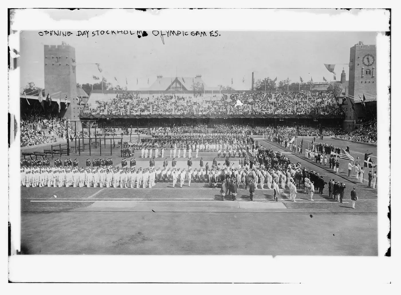 A hand-labelled glass-plate photograph of a packed Olympic stadium, with ranks of white-clad athletes standing in formation on the field before a grandstand full of spectators.