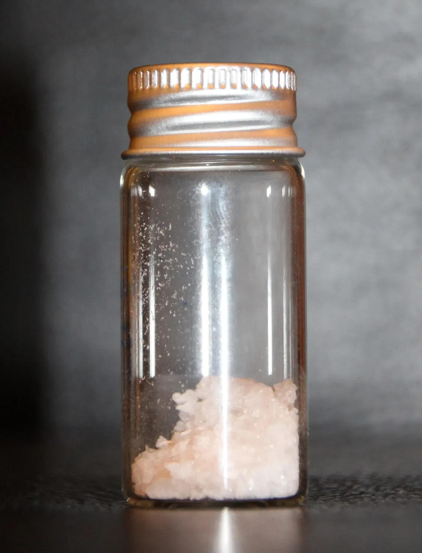 A small glass specimen jar with a screw cap, containing a cluster of small, pinkish-white crystals in its bottom corner.