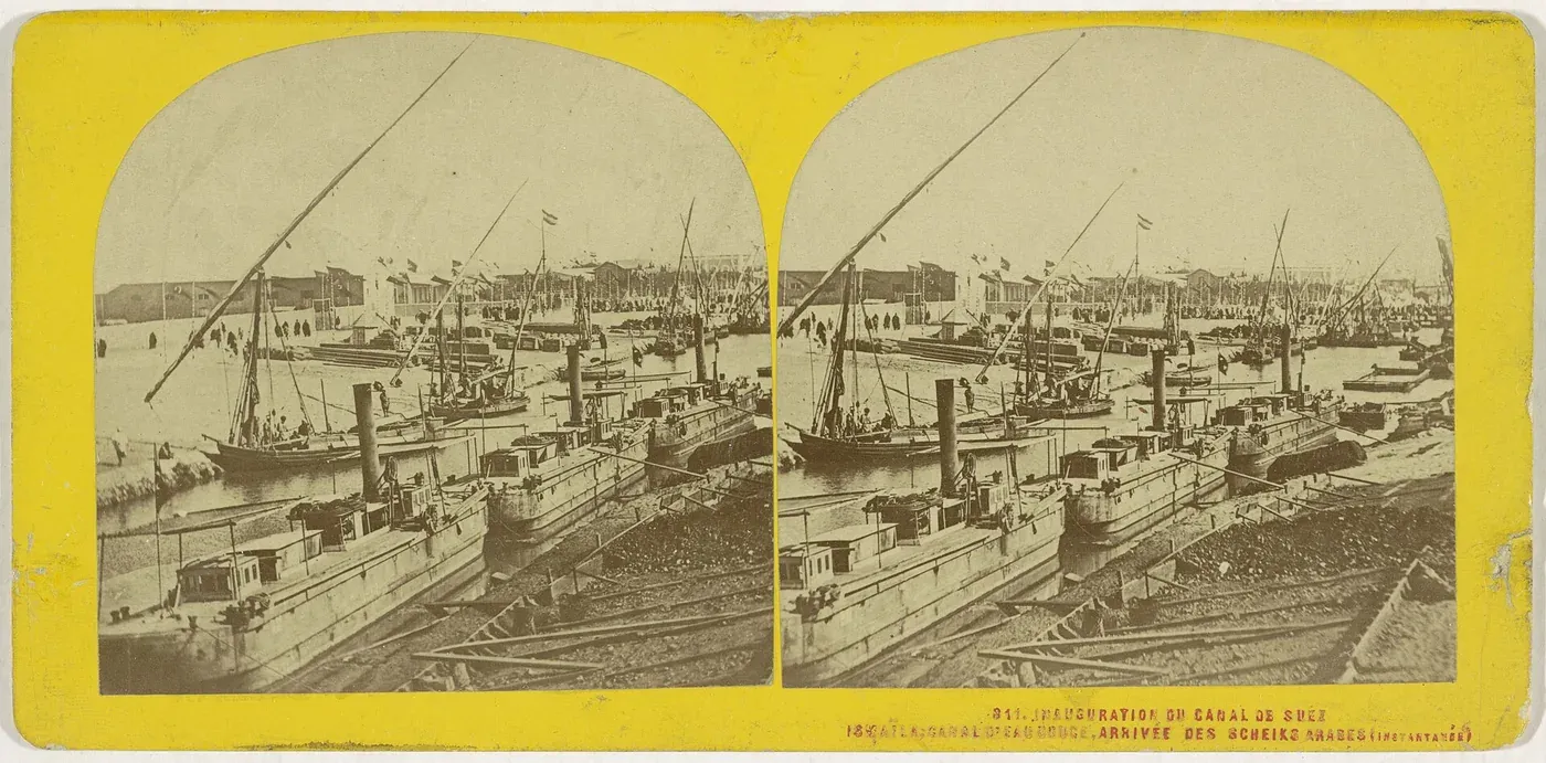 A stereograph photograph of small steam-powered boats and barges moored along a narrow desert waterway lined with low buildings and ships' masts.