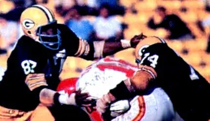 Green Bay Packers defensive linemen in dark jerseys tackle a Kansas City Chiefs player in white and red, mid-play at Super Bowl I.