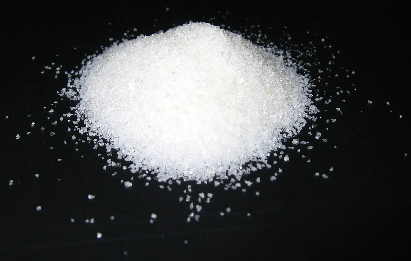 A close-up photograph of a small heap of coarse white crystalline granules scattered against a black background.