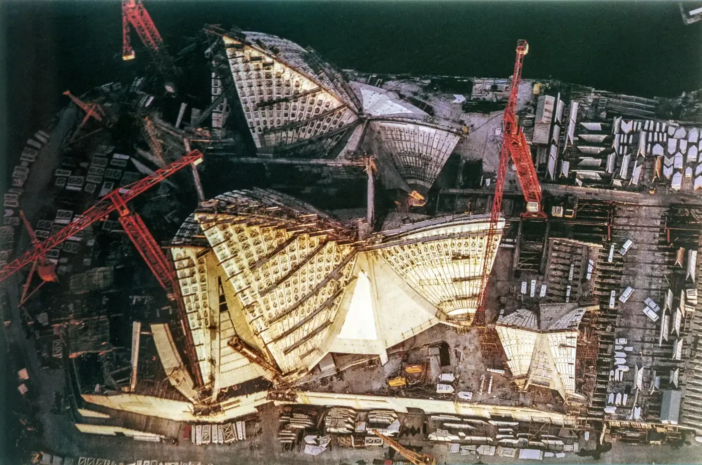 An aerial view of the Sydney Opera House under construction in 1965, showing the ribbed skeletal framework of the shell roofs partly clad in white tile panels, surrounded by tower cranes.