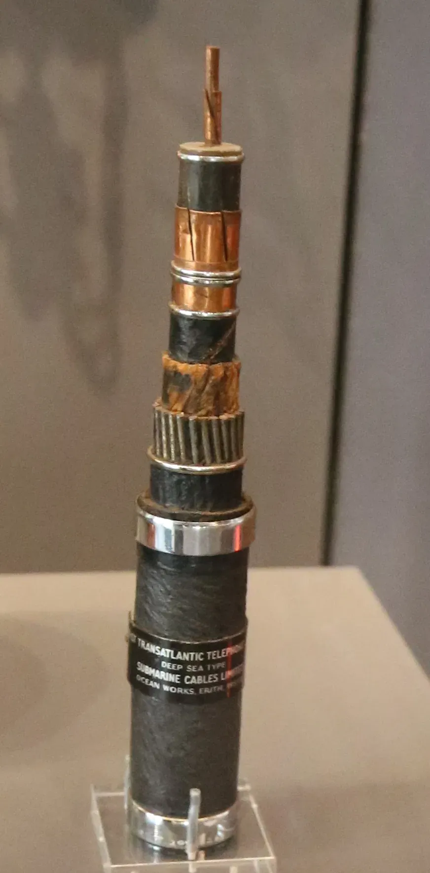 A museum display of a short vertical section of transatlantic telephone cable with its layers of coating progressively stripped back from top to bottom, exposing a central copper conductor, then successive layers of insulation, steel armour wires, and an outer wrapped jacket labelled TRANSATLANTIC TELEPHONE, DEEP SEA TYPE, SUBMARINE CABLES LIMITED.