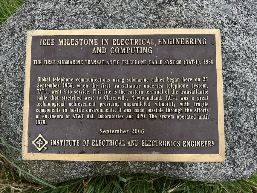 A bronze plaque mounted on a granite boulder, reading IEEE MILESTONE IN ELECTRICAL ENGINEERING AND COMPUTING, THE FIRST SUBMARINE TRANSATLANTIC TELEPHONE CABLE SYSTEM (TAT-1), 1956, with a paragraph of explanatory text below.