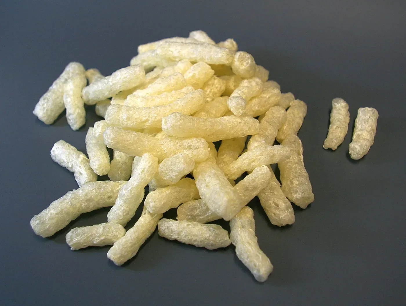 A small heap of pale yellow, cylindrical, foam-like packing chips on a dark background, with two loose pieces set apart from the pile.