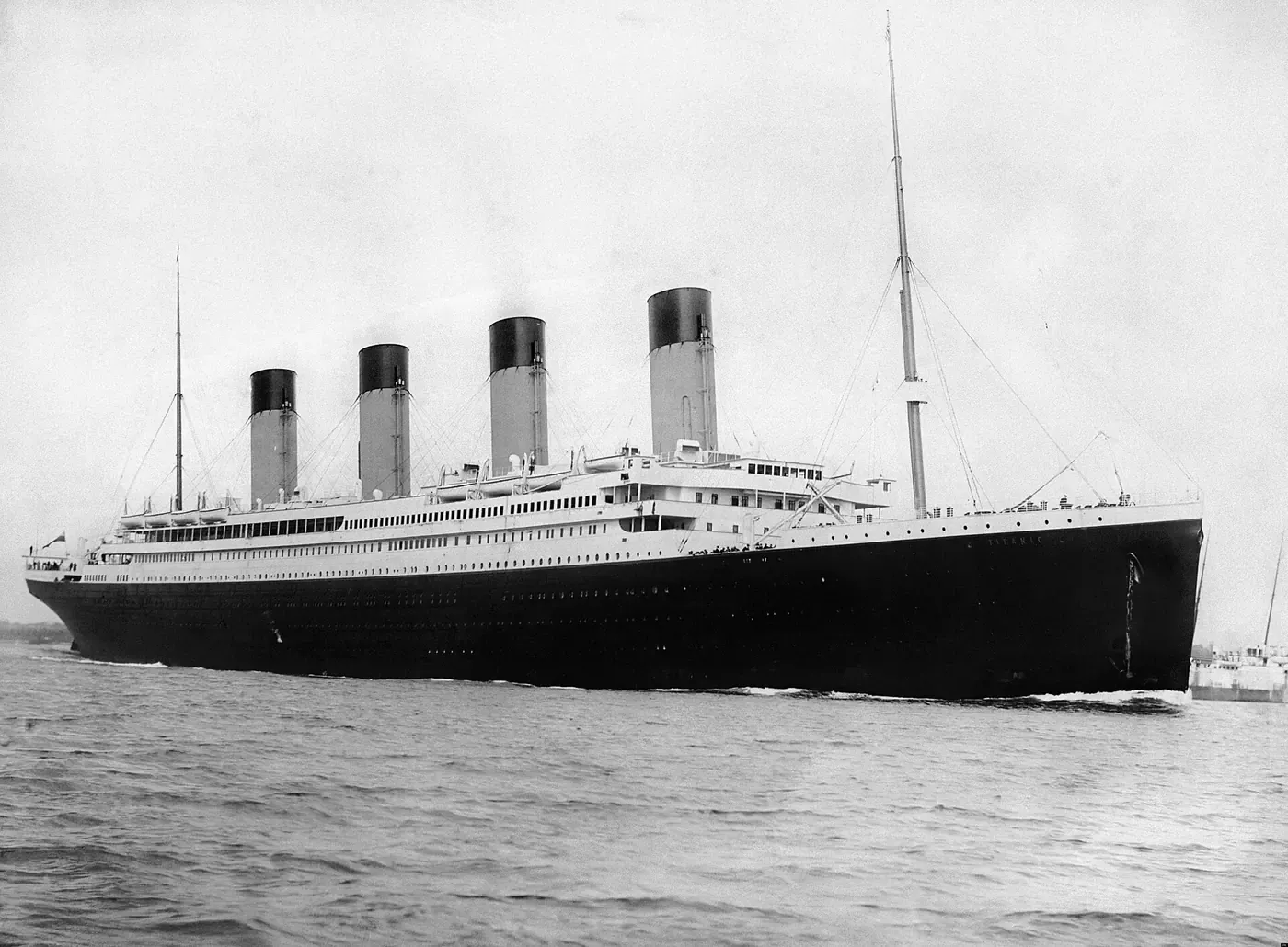 A large four-funnelled ocean liner steaming through calm water, smoke trailing from its stacks, with a smaller vessel visible near its stern.