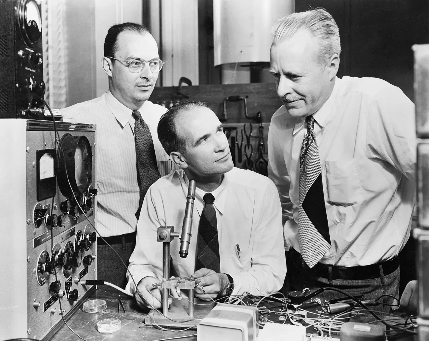 A black-and-white photograph of three men in shirtsleeves and ties gathered around a laboratory bench crowded with electronic test equipment, dials, wires and a small mounted device, with one man seated and two standing behind him.