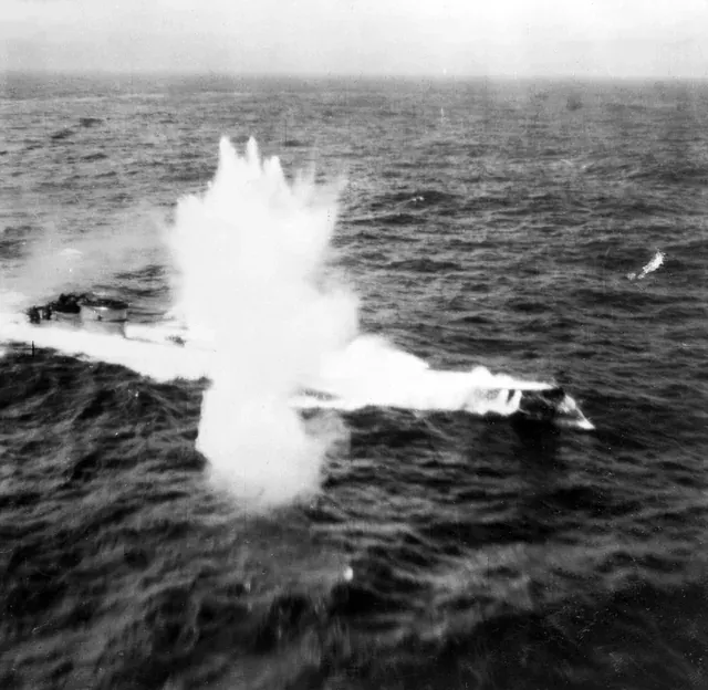 An aerial view of the ocean surface with a German U-boat under attack, a large plume of white water and spray thrown up beside its hull from a depth-charge or bomb strike.
