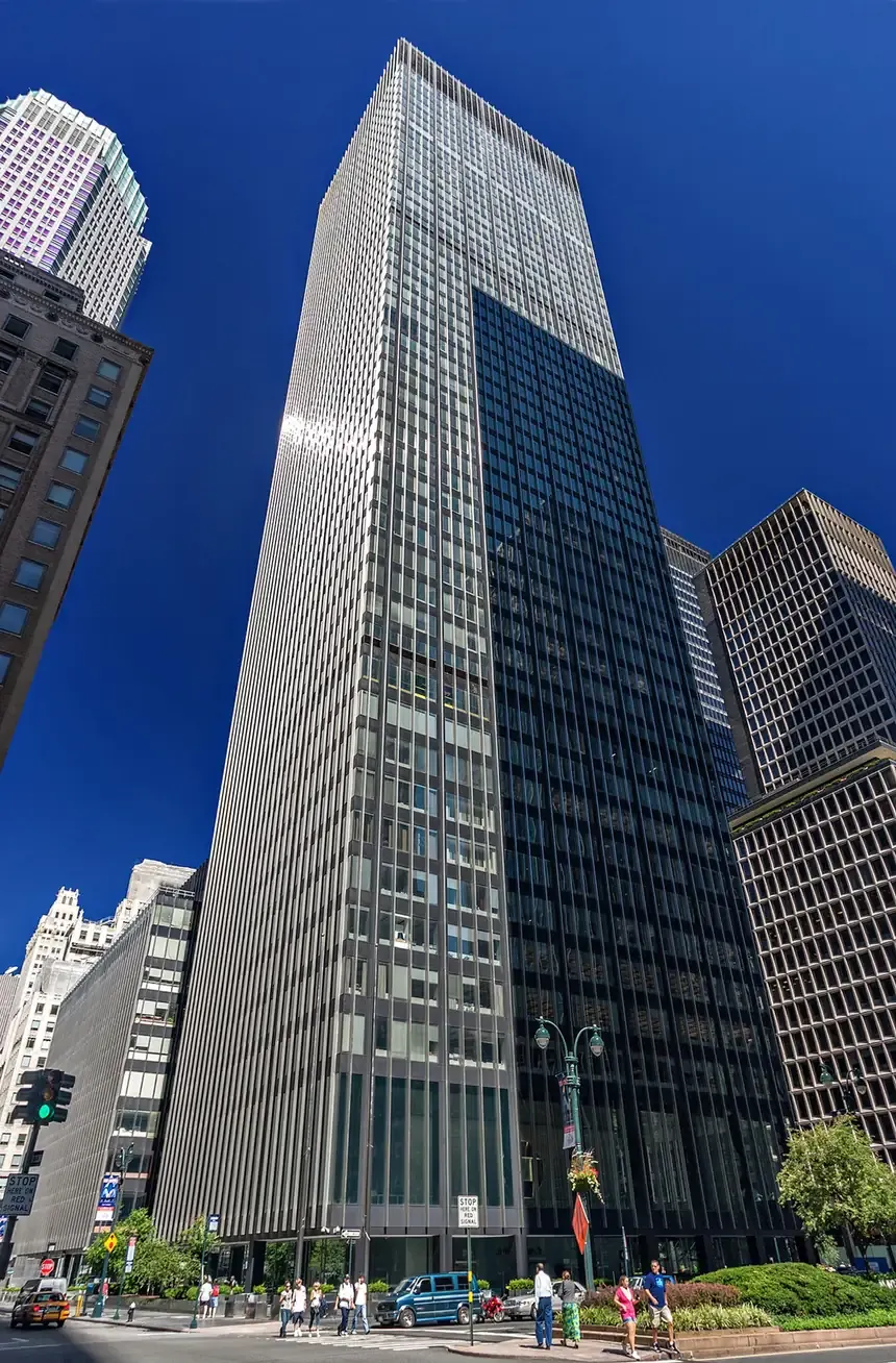 A very tall, narrow, glass-and-metal skyscraper viewed from street level looking straight up, its curtain-wall façade reflecting the sky, flanked by older stone buildings.