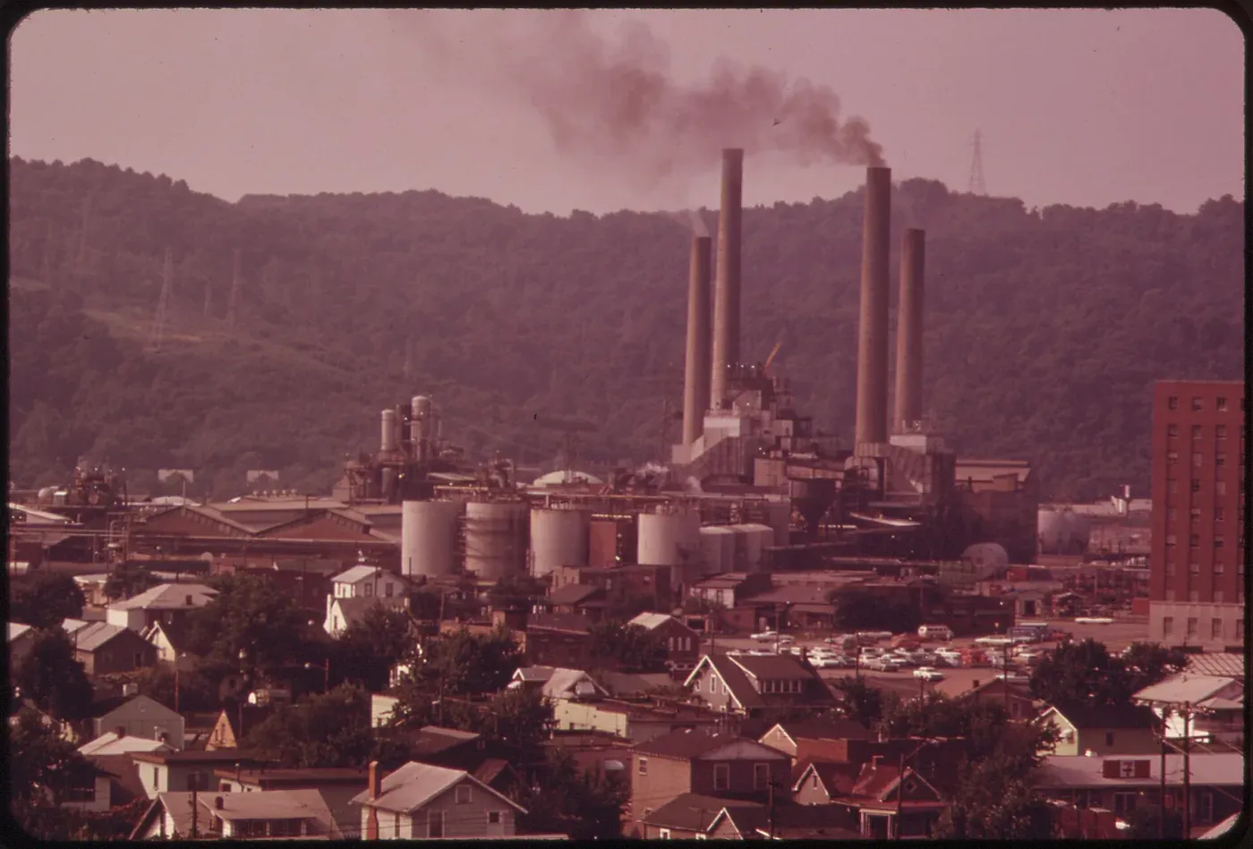 A hazy, sepia-toned view over a river town toward a large chemical plant with five tall smokestacks, one venting dark smoke, with storage tanks and rows of houses in the foreground.
