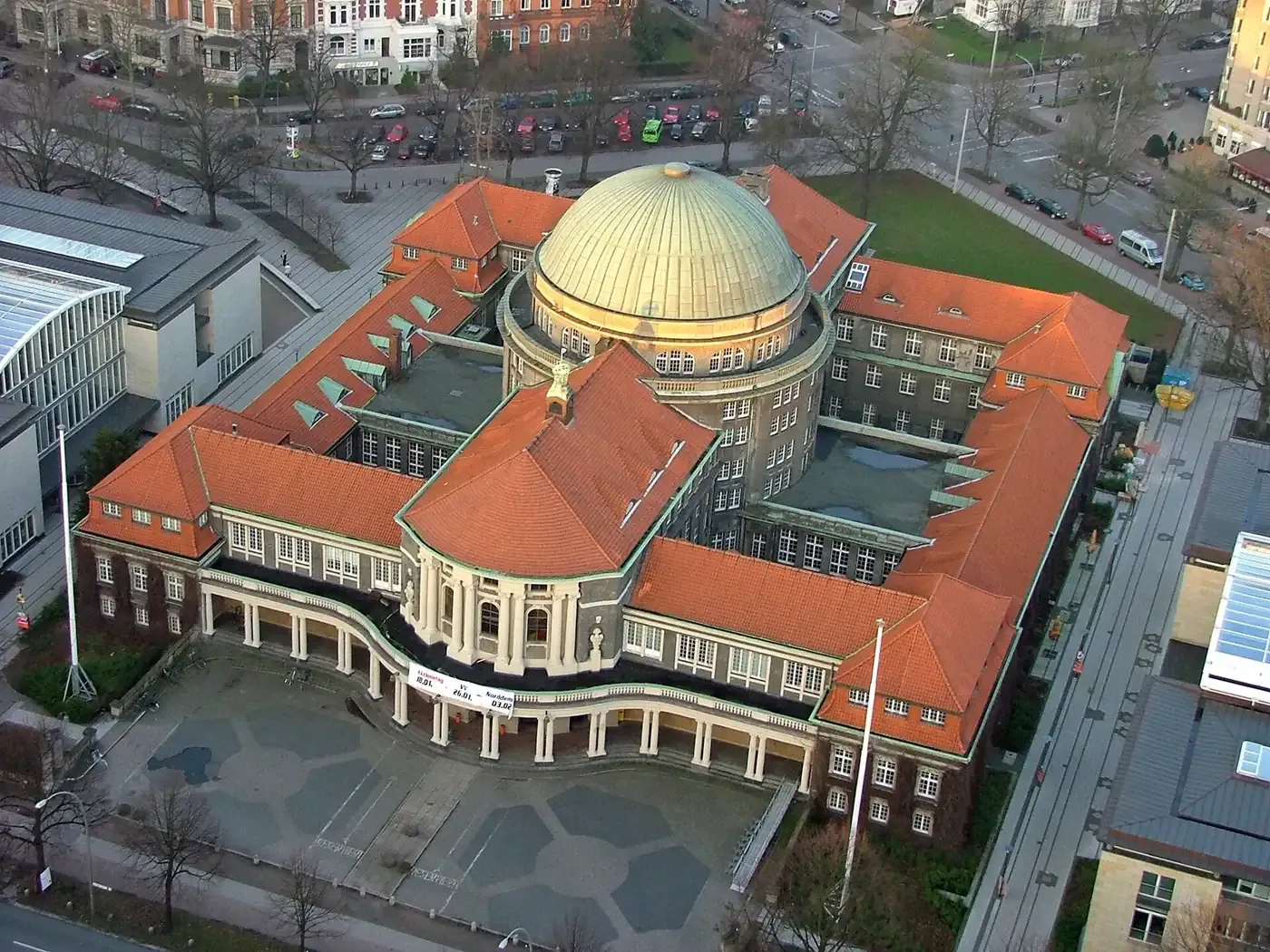 An aerial view of an ornate early-twentieth-century domed building with red tile roofs, forming a large enclosed courtyard in a European city centre.