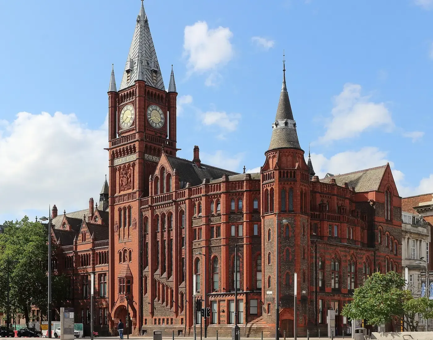 A red-brick and terracotta Victorian Gothic building with a tall clock tower topped by a spire, set against a blue sky with scattered clouds.